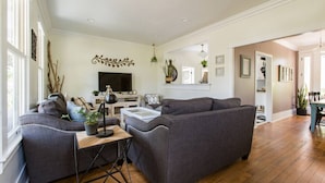 A spacious living room with a large gray sectional sofa, a coffee table, and a TV. There is a dining area with a table and chairs visible in the background. - Iconic Modern Farmhouse+walkable+views+BBQ (San Luis Obispo)