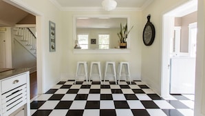A hallway with a checkered floor, a staircase, a white wall with a clock, and a window with plants. - Iconic Modern Farmhouse+walkable+views+BBQ (San Luis Obispo)