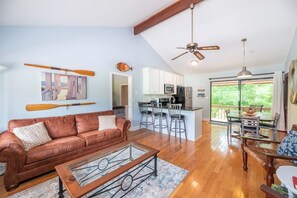 A spacious living area with a brown leather sofa, a coffee table, and a dining area with a table and chairs. A kitchen with white cabinets and a microwave is visible in the background.