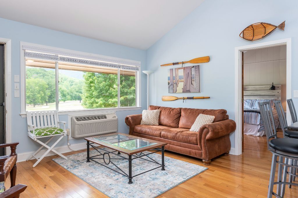 A living room with a brown leather sofa, a glass coffee table, a white folding chair, and a large window with a view of trees.