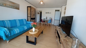 Een woonkamer met een blauwe bank, een houten salontafel met een bloevas, een eethoek met een tafel en stoelen, een televisie en een koelkast.