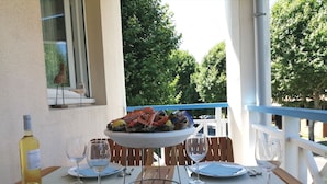 Een eettafel gedekt voor twee personen, compleet met borden, bestek en wijnglazen. Op tafel staat een schaal met zeevruchten, en er staat een fles wijn. De setting is buiten, met uitzicht op bomen en een hekwerk.
