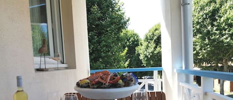 A dining table set for two with plates, cutlery, and wine glasses. A seafood platter is on the table, and a bottle of wine is visible. The setting is outdoors with a view of trees and a railing.