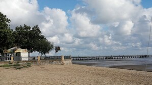 Een strand met een pier die het water in reikt, een omheind gebied met een klein gebouw, en bomen op de achtergrond.