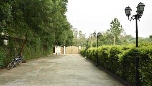 A paved pathway leading to a building with a small entrance, surrounded by dense greenery and a parked motorcycle. - Remarkable 6-bedroom Farmhouse in South Delhi (New Delhi)