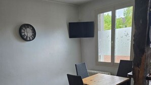 Een kamer met een houten eettafel, twee stoelen, een donkere bank, een wandklok en een televisietoestel.
