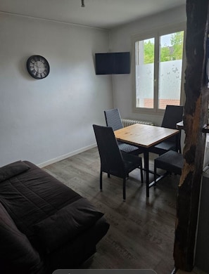 Ein Zimmer mit einem Holztisch, zwei Stühlen, einem dunklen Sofa, einer Wanduhr und einem Fernseher.