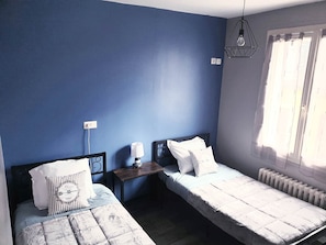 Ein Zimmer mit zwei Betten, einem Nachttisch mit Lampe, einer Pendelleuchte und einem Fenster mit Vorhängen.
