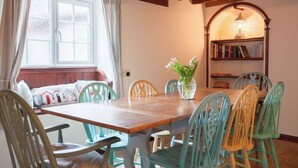 A dining area with a wooden table and chairs, a bookshelf, and a window with curtains. - Cosy Norfolk Cottage (King's Lynn)