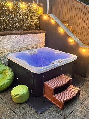 A hot tub with a wooden bench and two green cushions, surrounded by a wooden fence and string lights. - North Lodge (Lancaster)