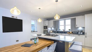 A modern kitchen with a central island, white cabinetry, pendant lighting, and a flat-screen TV mounted on the wall.