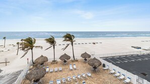 A beach view with lounge chairs, palm trees, and a parking area. - Luxury Beach Front Corner Beach Place #306 (Madeira Beach)