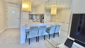 A modern kitchen with a central island, a dining area with four chairs, and a large flat-screen TV on a stand. - Fashion Avenue Dubai Mall Residences - Studio with balcony (Dubai)