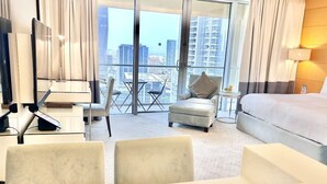 A hotel room with a bed, a desk with a chair, a small sofa, and a view of the city through large windows. - Fashion Avenue Dubai Mall Residences - Studio with balcony (Dubai)