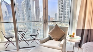 A modern hotel room with a large window offering a city view, a comfortable sofa, a small round table with a drink, and a folding chair. - Fashion Avenue Dubai Mall Residences - Studio with balcony (Dubai)
