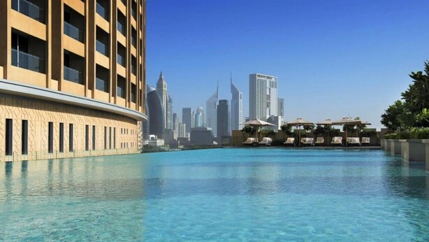 A swimming pool with a city skyline in the background, a building with a beige facade, and umbrellas on the poolside. - Fashion Avenue Dubai Mall Residences - Studio with balcony (Dubai)