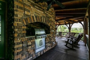An outdoor brick fireplace with a grill, wooden deck, and rocking chairs.