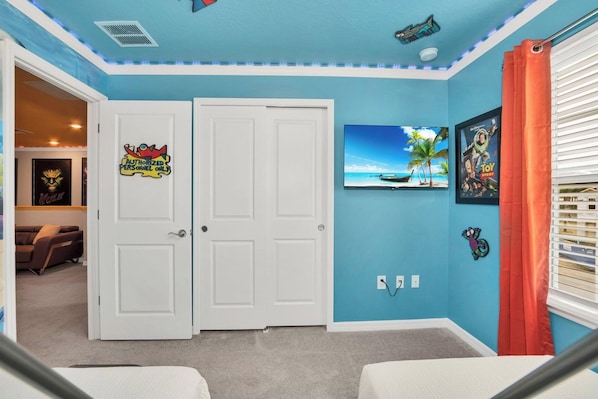 Una habitación con dos puertas blancas, techo con detalles azules, un televisor en la pared y una ventana con persianas.