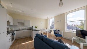 A modern kitchen with white cabinets, a dining area with a glass table, and a large window offering a view of the city. - Apartment 5 Isabella House Hereford (Hereford)