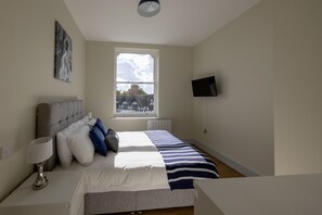 Ein Schlafzimmer mit einem Bett, einem Nachttisch mit Lampe, einem an der Wand montierten Fernseher und einem Fenster mit Blick auf ein Gebäude.