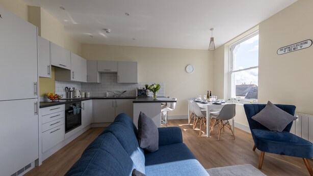 A modern kitchen with white cabinets, a dining area with a table and chairs, and a blue sofa in a living room. - Apartment 5 Isabella House Hereford (Hereford)