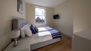 A bedroom with a bed, a nightstand, a lamp, a painting, a window, and a TV. - Apartment 5 Isabella House Hereford (Hereford)