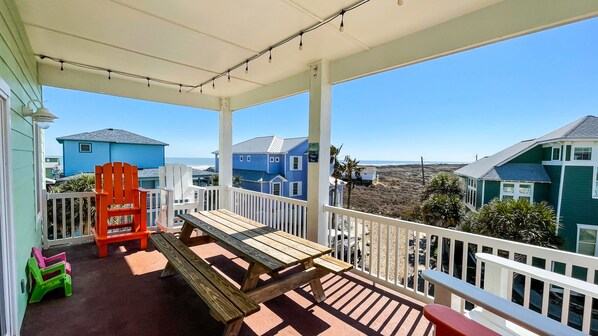 A covered outdoor area with a wooden bench and chairs, a white railing, and a view of blue houses and the beach.