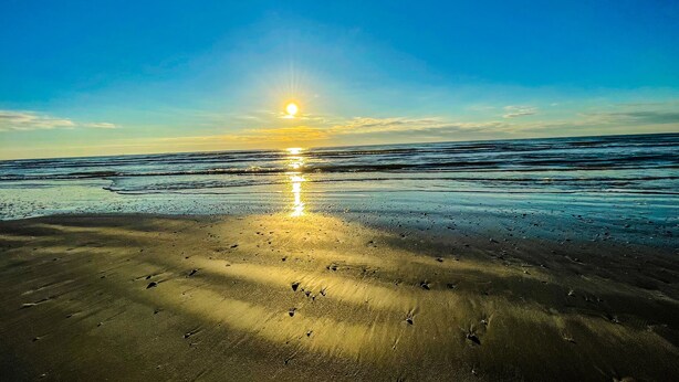 Ein Strand mit Sonnenuntergang, dessen Licht sich im feuchten Sand spiegelt und einen Lichtweg bis zum Horizont bildet.