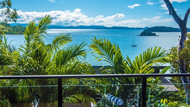 Une vue depuis un balcon donnant sur une mer calme, avec une petite île et un voilier visibles au loin.