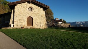 A stone building with a wooden door and a circular window, surrounded by a grassy area and a paved path. - House La Galinette 9 people Heated swimming pool Hammam Jacuzzi Sauna (Izeaux)