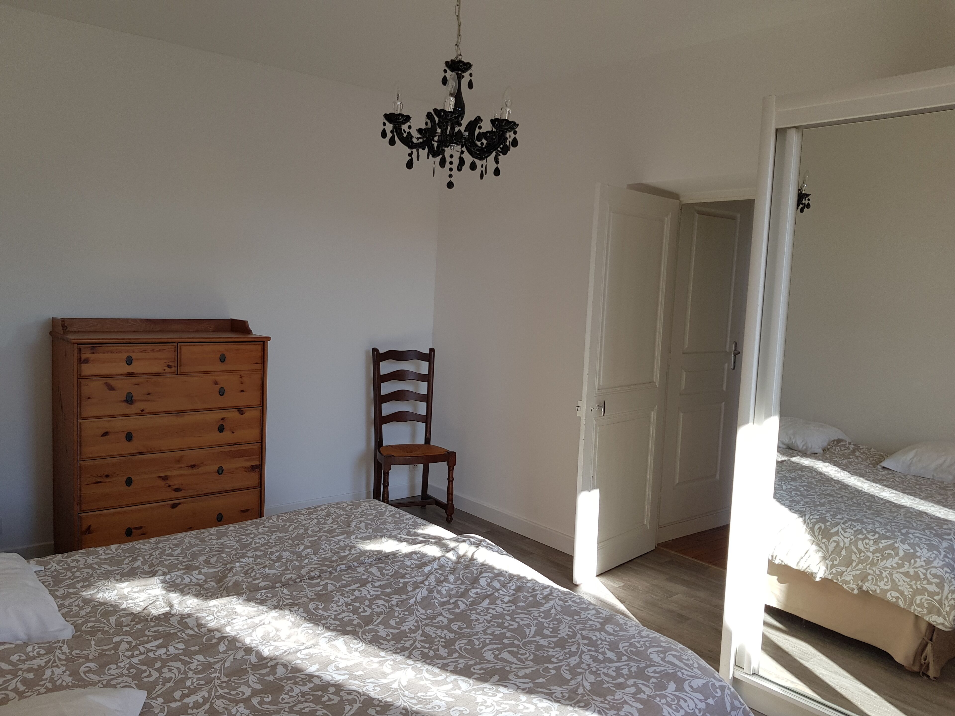 A bedroom with a wooden dresser, a chair, a bed with patterned bedding, a chandelier, and a door leading to another room.