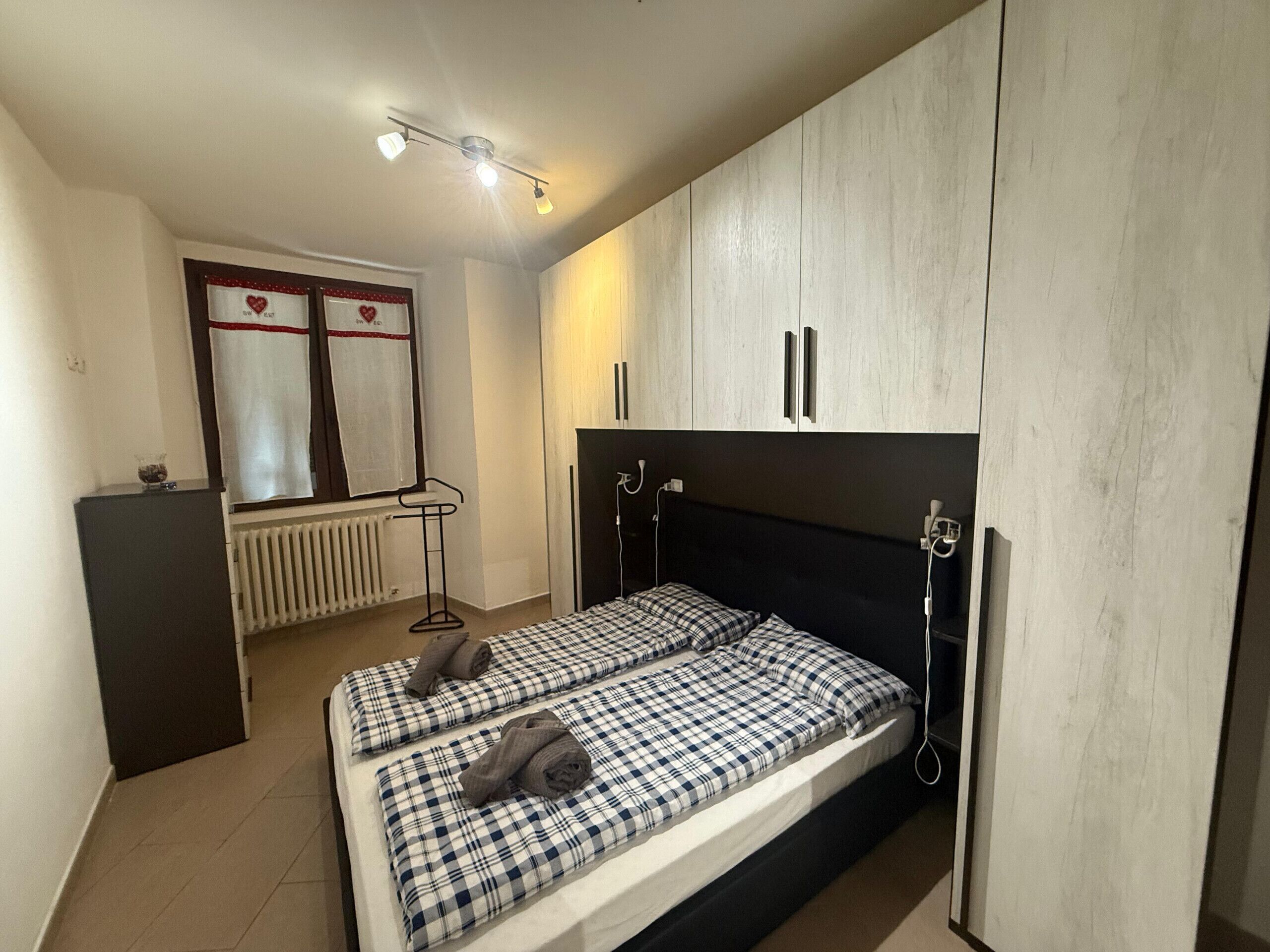 A bedroom with a bed, a radiator, a window with curtains, and a ceiling light fixture.