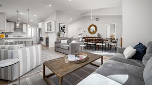 A modern living room with a grey sofa, striped armchair, and a wooden coffee table. There is a dining area with a table and chairs, and a kitchen in the background.