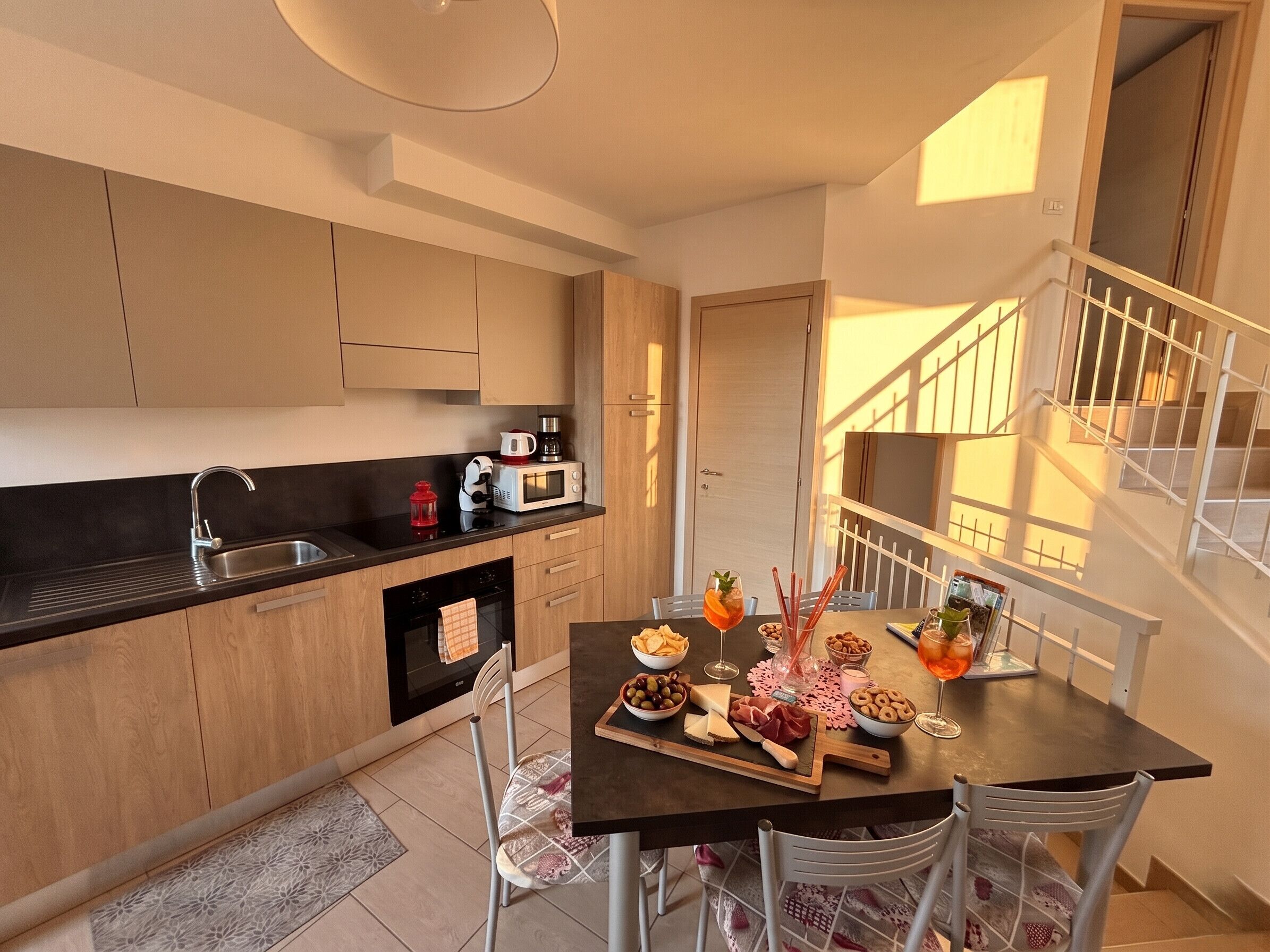 A modern kitchen with a dining area, wooden cabinets, a black countertop, and a staircase with a railing.