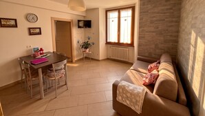 Una sala de estar con un sofá, una mesa de comedor con sillas, un televisor montado en la pared y un ventanal con cortinas.