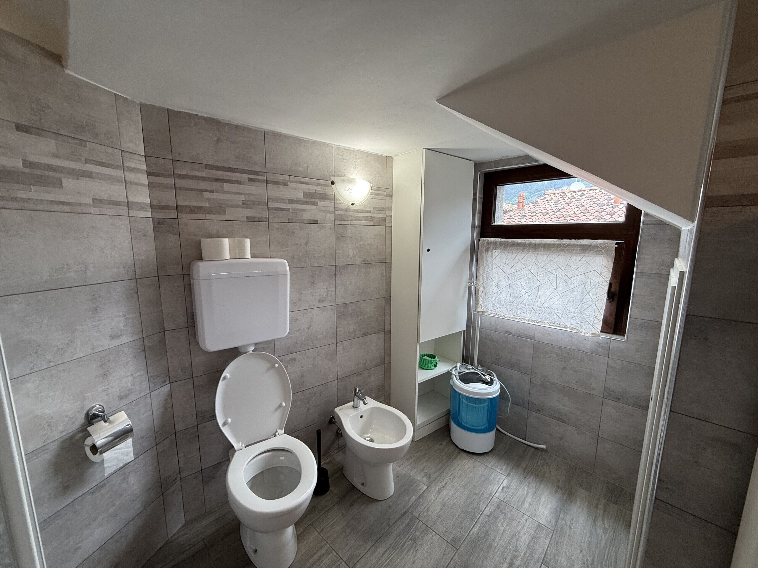 A bathroom with a toilet, bidet, and a window with a view of buildings.