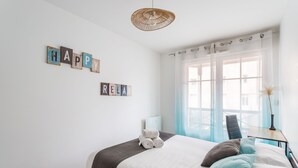 A neatly made bed with a white comforter, two rolled towels, and two pillows. There are two framed words on the wall, "HAPPY" and "RELAX". A wicker floor lamp hangs above the bed, and a window with sheer curtains is visible.