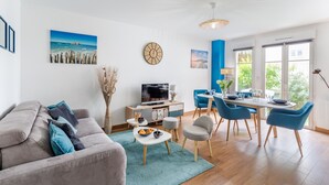 A modern living room with a grey sofa, a dining table with blue chairs, a TV, and artwork on the walls.