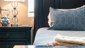 Uma cama bem arrumada com travesseiro estampado, luminária de cabeceira e um recipiente de vidro decorativo sobre um criado-mudo de madeira escura.