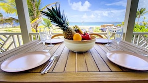 Una mesa con frutero, platos, cubiertos y vasos sobre una terraza de madera con vista a una playa.