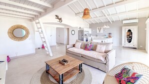 Amplia sala de estar con techo alto, una mesa de centro de madera, un sofá con cojines coloridos, un sillón de mimbre y un espejo redondo en la pared.