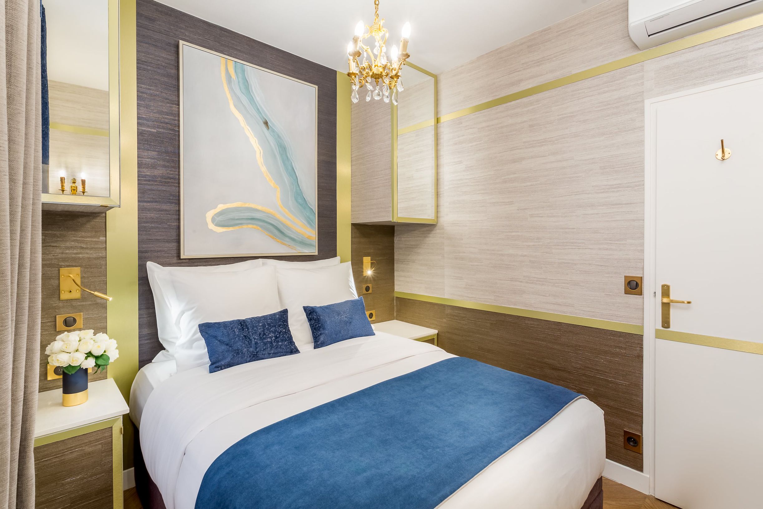 A neatly arranged hotel room with a large bed, a bedside table with a vase of flowers, a wall-mounted artwork, and a modern chandelier.