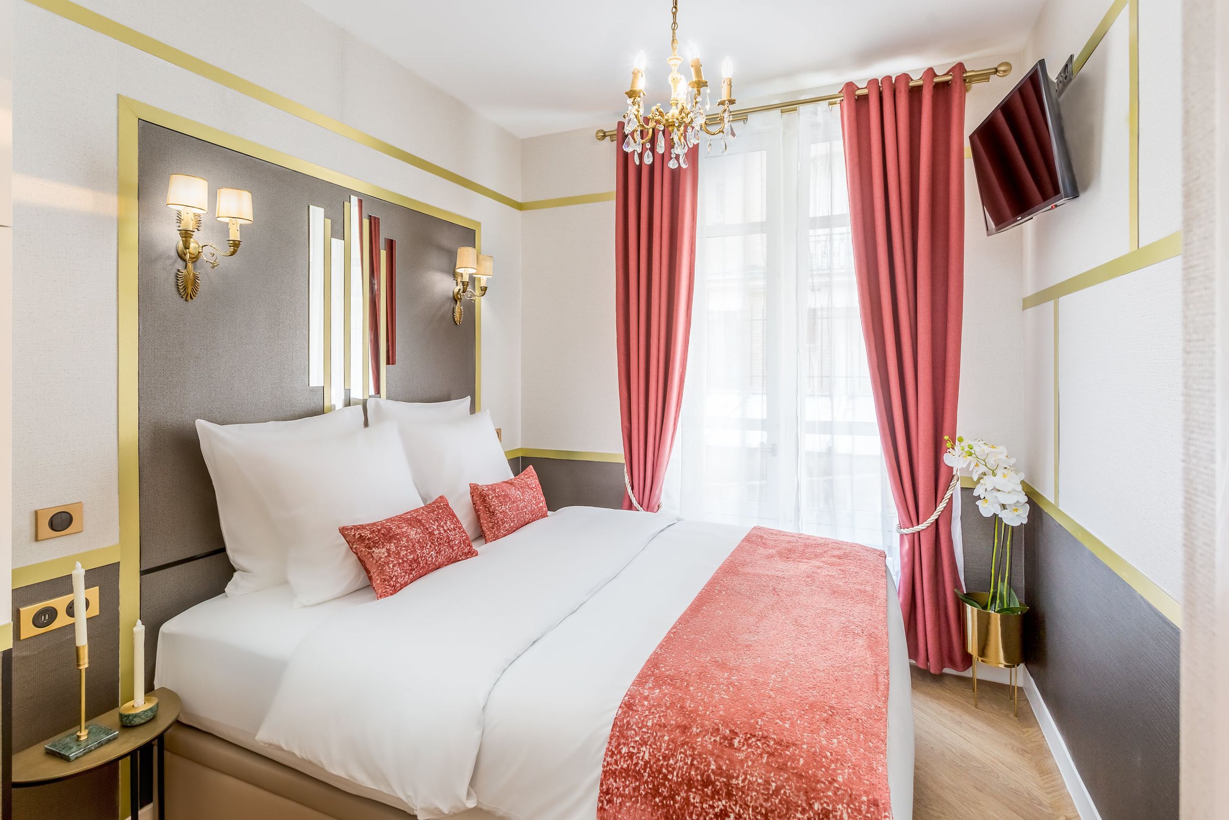 Hotellihuone, jossa on suuri sänky, koristeellinen kattokruunu, litteänäyttöinen televisio ja punaiset verhot.