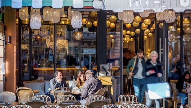 Un café avec des chaises et des tables en osier, un auvent à rayures et des guirlandes lumineuses décoratives suspendues.