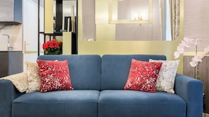 En blå soffa med rött och vitt mönstrade kuddar, en vas med röda blommor och en spegel med en guldfärgad ram.