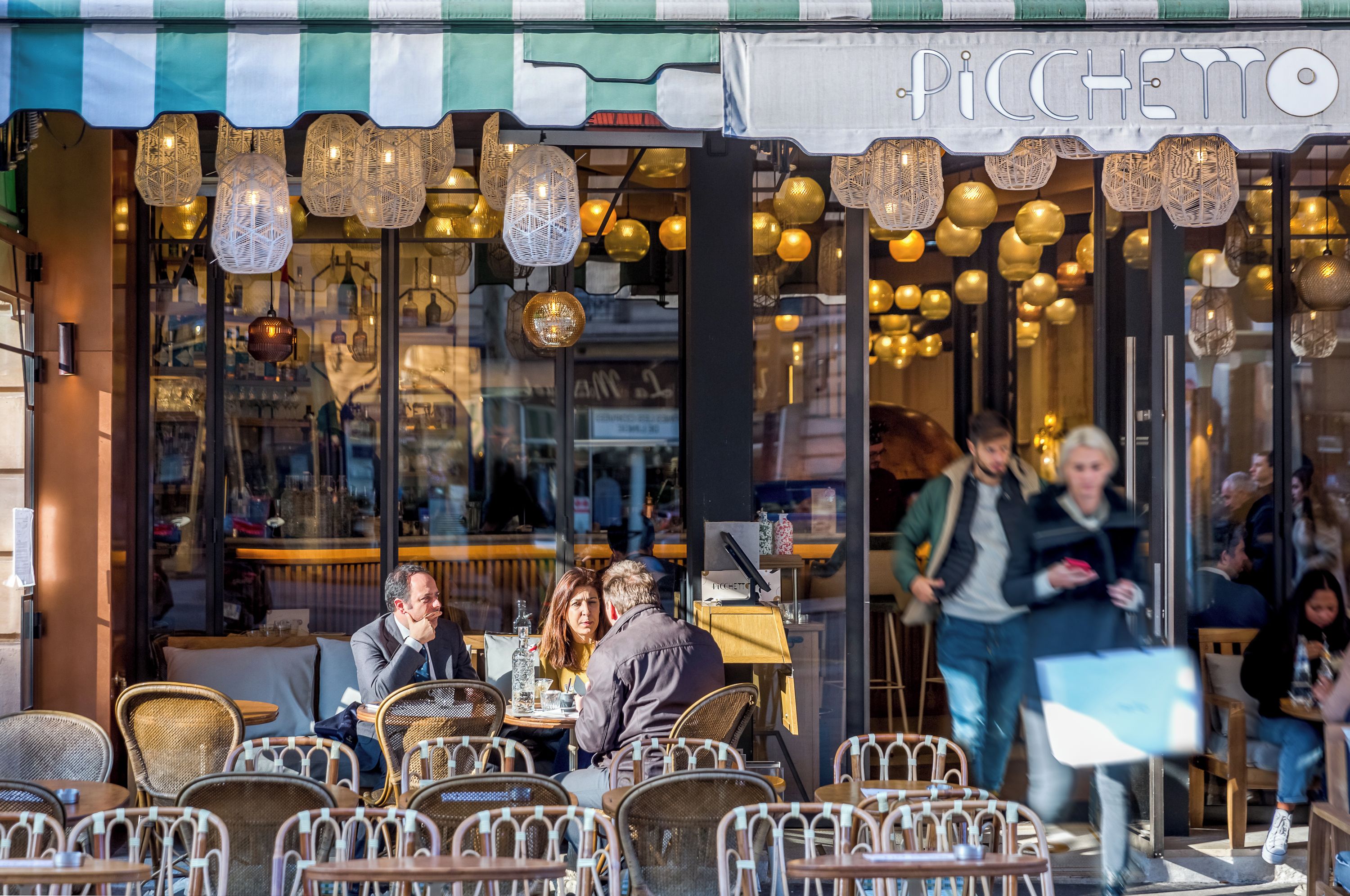 Un caffè all'aperto con sedie e tavoli in vimini, un tendone a righe e luci decorative appese.