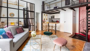 A spacious living area with a sofa, ottoman, and coffee table, a kitchen with bar stools, and a staircase leading to an upper level.