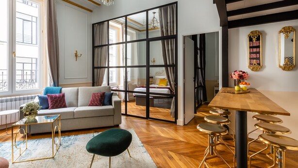 A modern living room with a sofa, a coffee table, a dining table with stools, and a large mirror.
