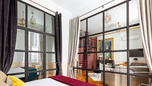 A modern hotel room with a large bed, a sofa, and a view of a cityscape through floor-to-ceiling windows.