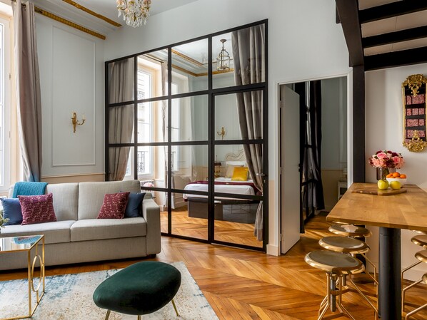 A modern living room with a sofa, a coffee table, a dining area with stools, and a large window with curtains.